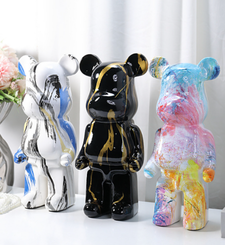 Three glossy bear-shaped decorative figures displayed side by side on a white surface against a soft white curtain background. The left figure is white with blue and gray paint-stroke patterns, the center figure is black with gold paint-stroke patterns, and the right figure is multicolored with pink, blue, yellow, and orange paint-splatter patterns.
