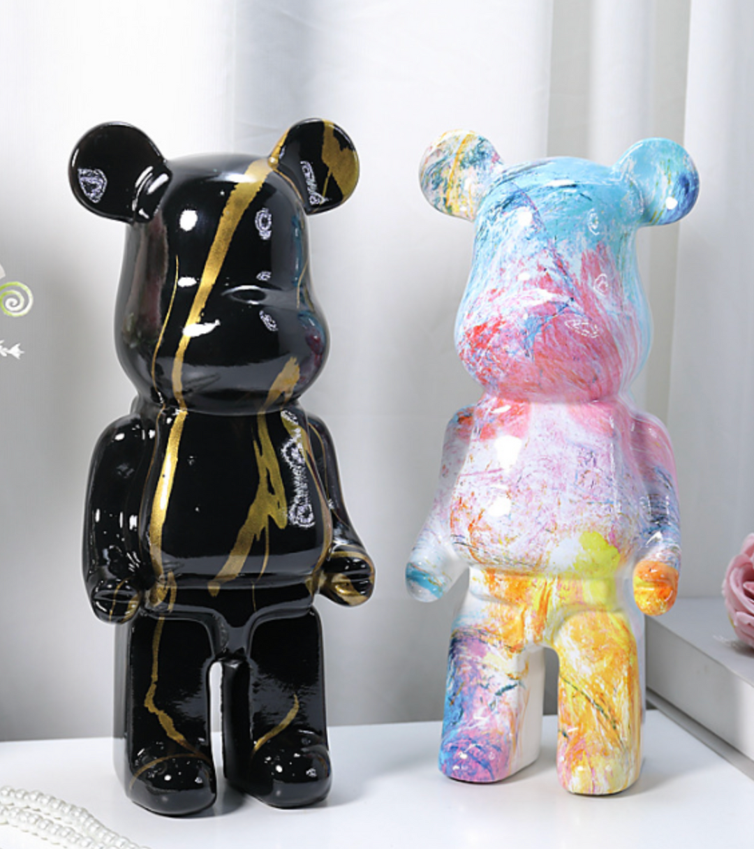 Two bear-shaped decorative figurines displayed side by side on a white surface against a soft white curtain background. The left figurine is glossy black with metallic gold marbled streaks, and the right figurine has a multicolor paint-splatter finish with blue, pink, yellow, and white tones.