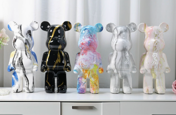 Five Bearbrick-style collectible figures lined up on a white surface against a soft white curtain background. The figures have glossy marbled and paint-splatter finishes in various colorways: white with blue and black marbling, black with gold veining, blue/pink/purple splatter, clear/gray marbling, and pink/white/gold marbling.