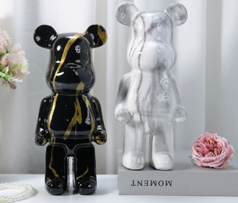 Two bear-shaped decorative figurines displayed side by side on a shelf: one glossy black with gold marbling and one translucent white with gray marbling. A pink flower and a small pearl strand sit beside them, with the words 'MOMENT' visible on the base.