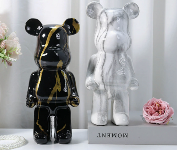 Two bear-shaped decorative figurines displayed side by side on a shelf: one glossy black with gold marbling and one translucent white with gray marbling. A pink flower and a small pearl strand sit beside them, with the words 'MOMENT' visible on the base.